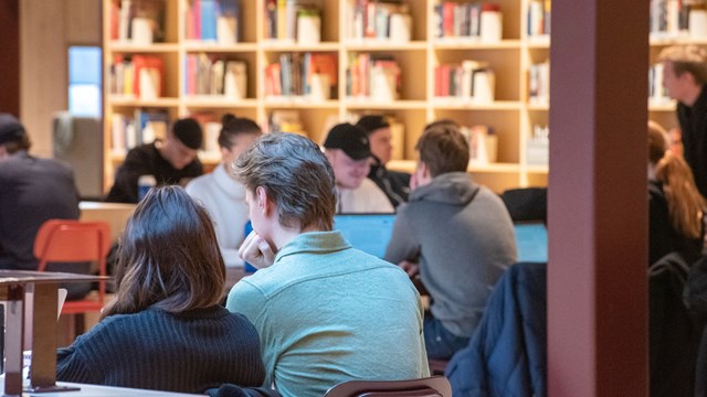 Students sitting at a number of tables with a book shelf behind them.
