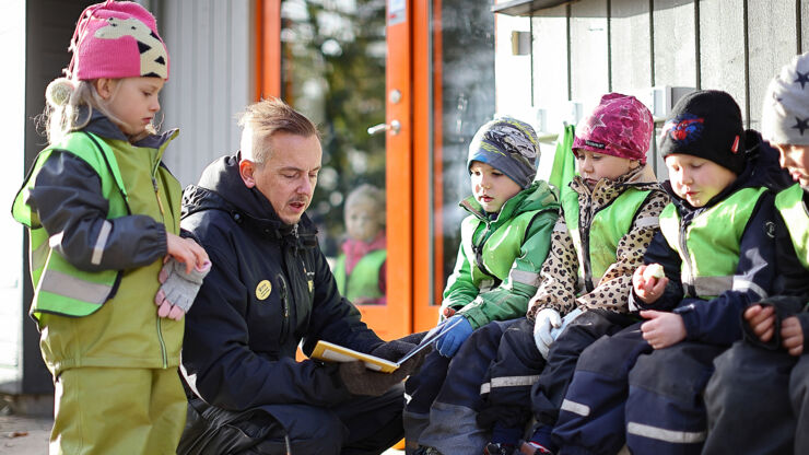 En lärare sitter på huk utomhus och håller i en bok. Runt läraren sitter barn i vinterkläder och lyssnar till läraren. 