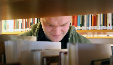 Jim Blomqvist läser, sedd genom bokhyllorna
