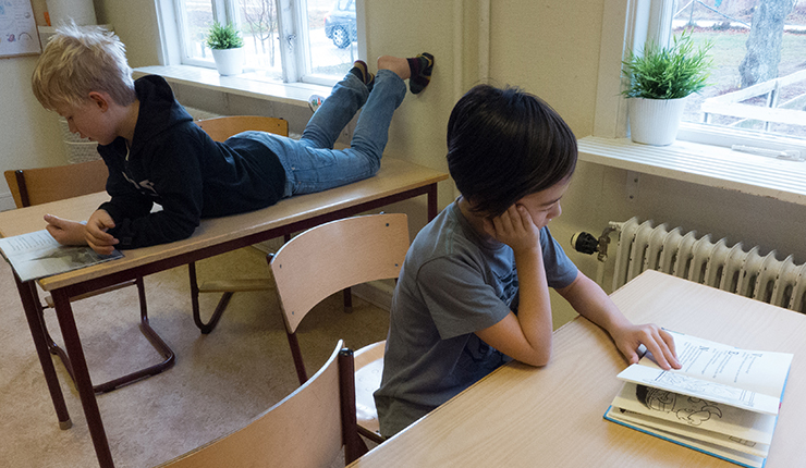 Två läsande elever i klassrum. En av dem ligger på borden med fötterna mot väggen.