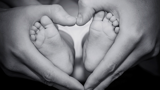 Mamma håller om en babys fötter. Hennes händer är formade som ett hjärta. / Mother holding the feet of a baby. Her hands make a shape of a heart.