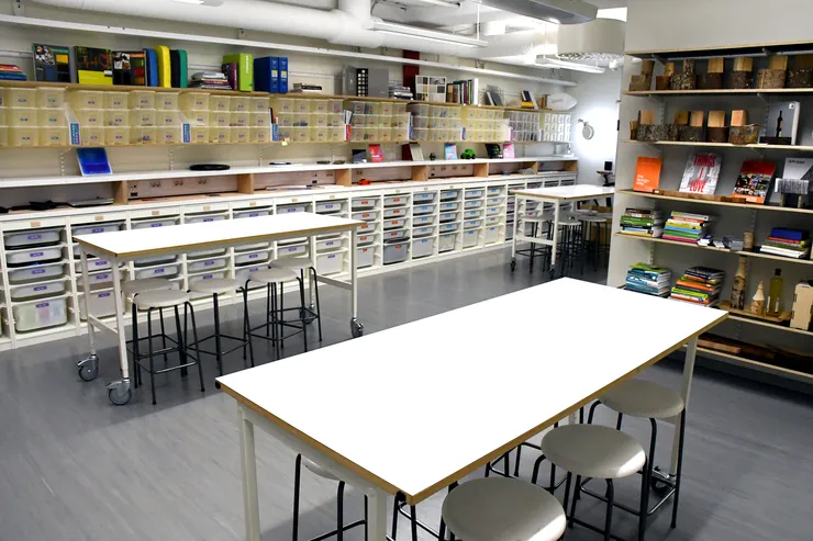 Material Realisation Laboratory. In the empty room there are three tables, as well as stools for these. One of the walls is largely filled with plastic boxes.