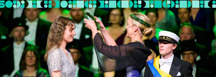 A woman puts a laurel wreath on another woman's head. People in formal wear in the background.