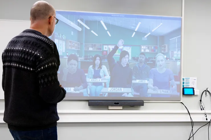 A teacher stands in front of a screen with digital students.