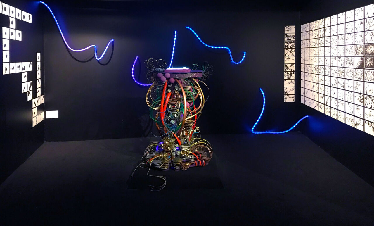Art installation in a dark room featuring a central sculptural piece of tangled wires and glowing elements, with abstract black-and-white artworks displayed on the surrounding walls.