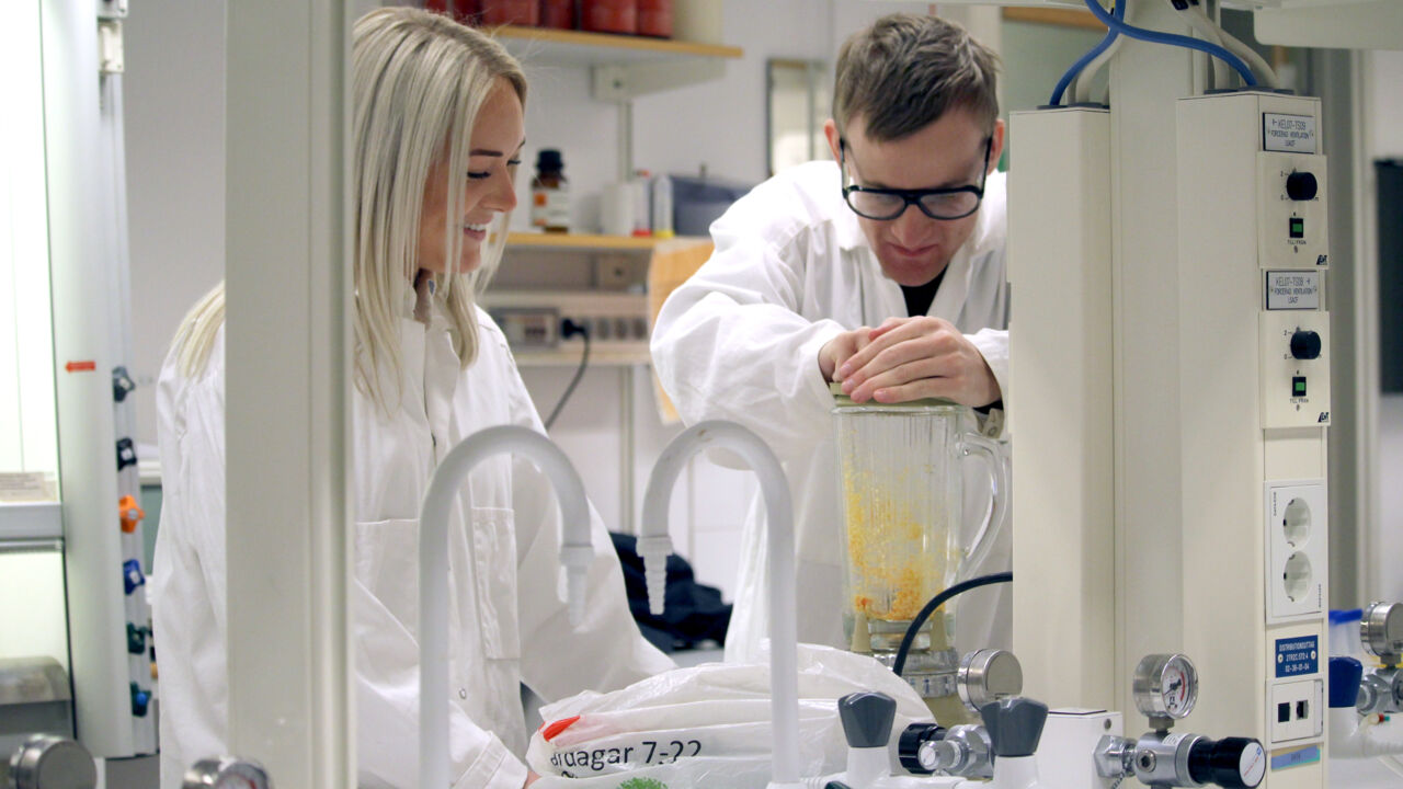En man och kvinna i labbmiljö. Locket till en mixer hålls nere medan vätskor blandas.