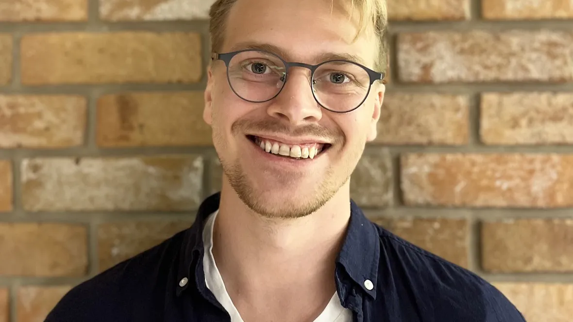 Leende blond man med glasögon. Gul tegelvägg i bakgrunden