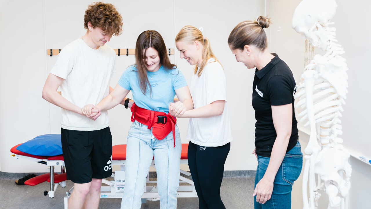 Tre fysioterapeutstudenter övar uppresning av patient med midjebälte. En lärare tittar på. 