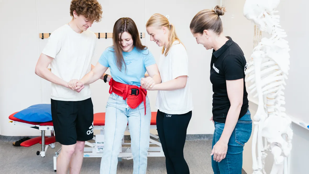 Tre fysioterapeutstudenter övar uppresning av patient med midjebälte. En lärare tittar på. 