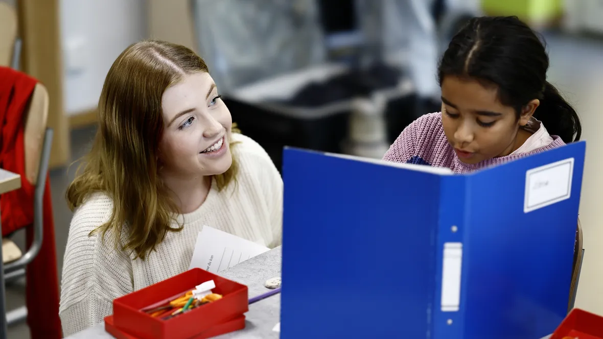 Kvinnlig lågstadielärare sitter vid en elevs bänk och hjälper till med en uppgift.
