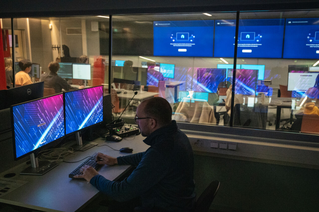 Large room with students at computers and a smaller control room with a course leader at the computer