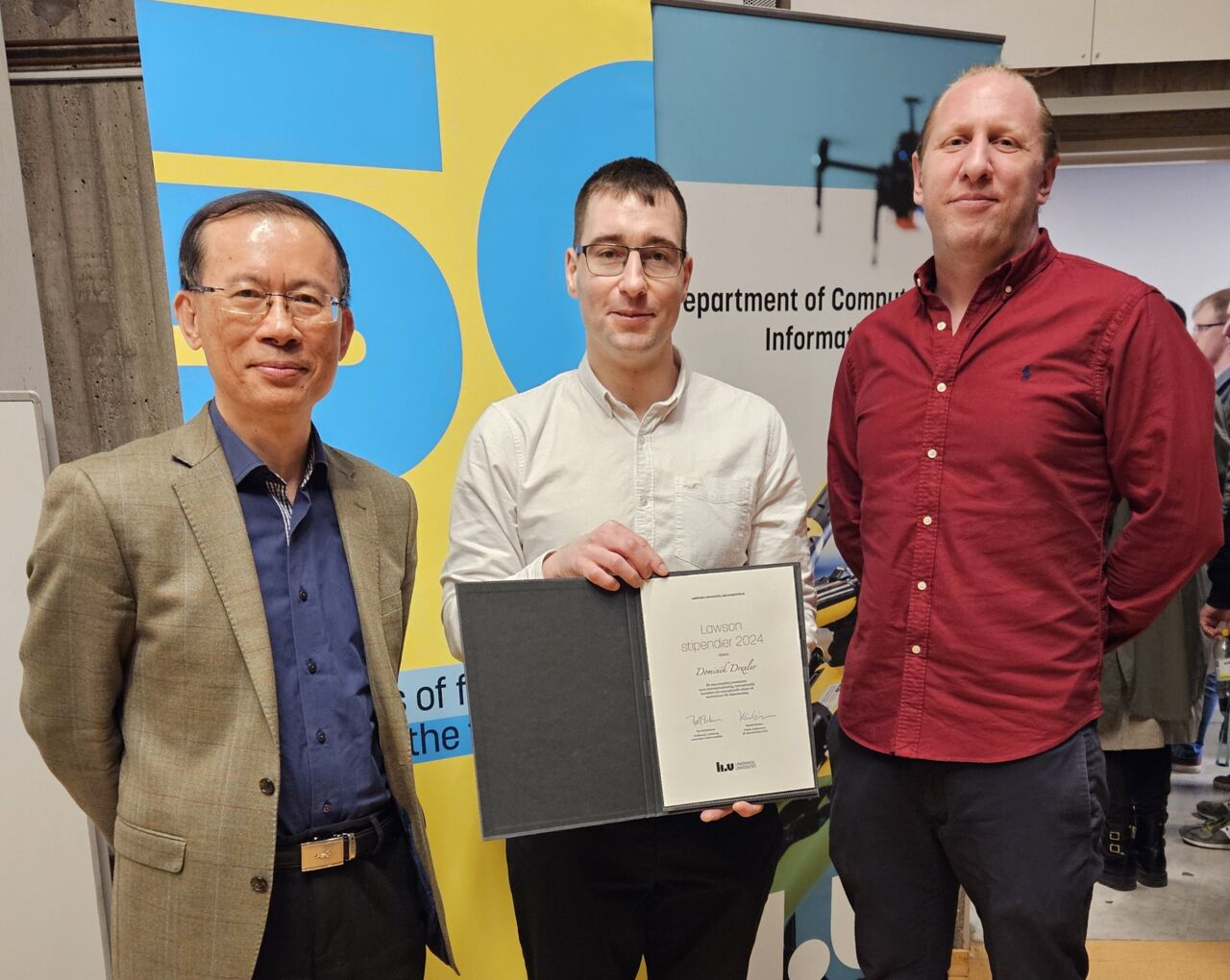 Tre män som står upp, den i mitten visar upp ett diplom