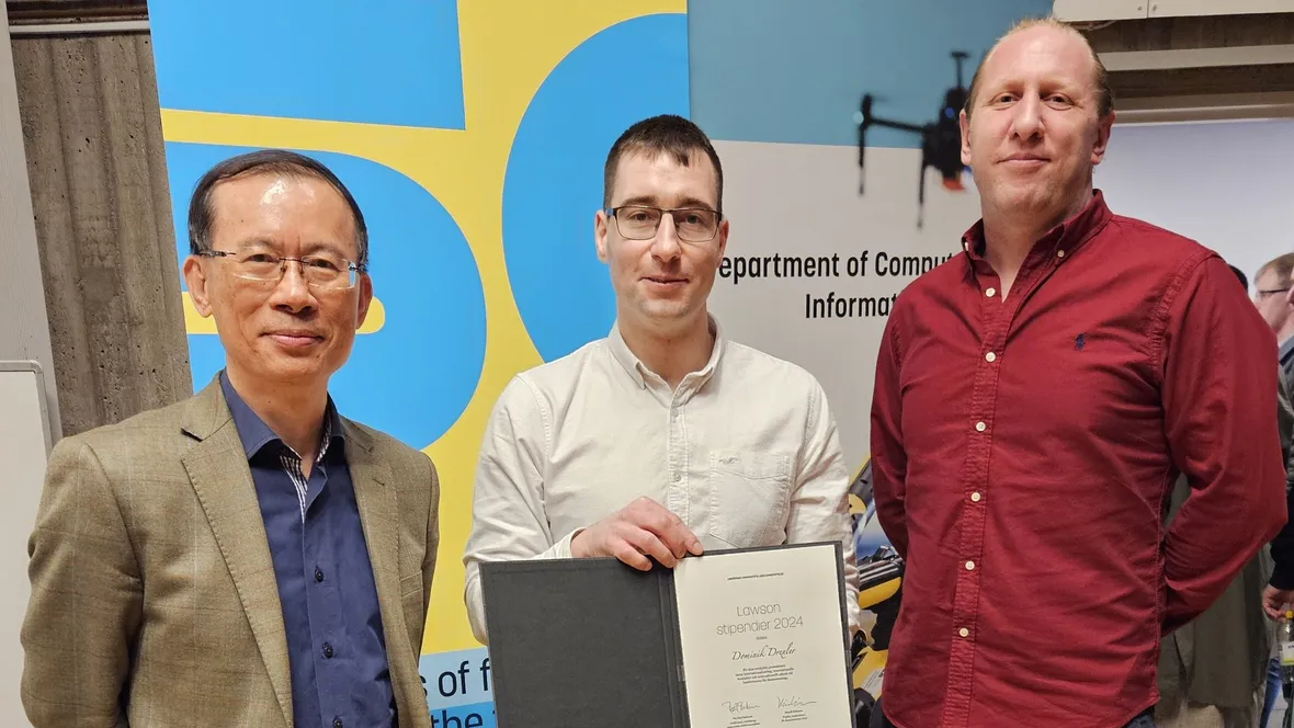 Three men standing up, the one in the middle is holding up a diploma