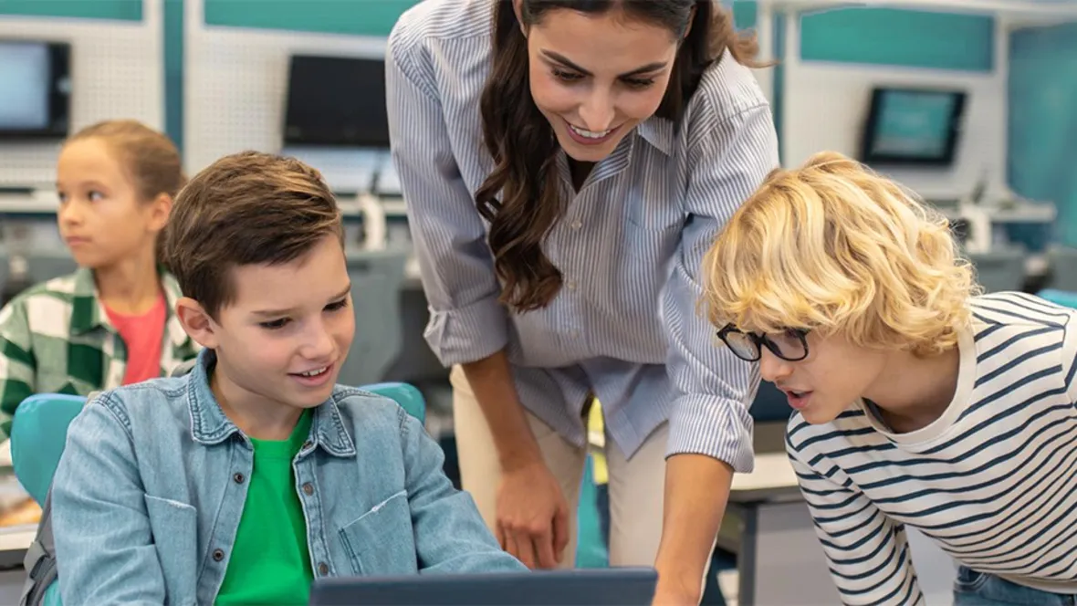 Teacher and children in classroom in front of screen. Computers in the background.