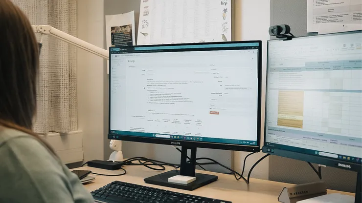 Två datorskärmar står på ett skrivbord, en person använder datorn.