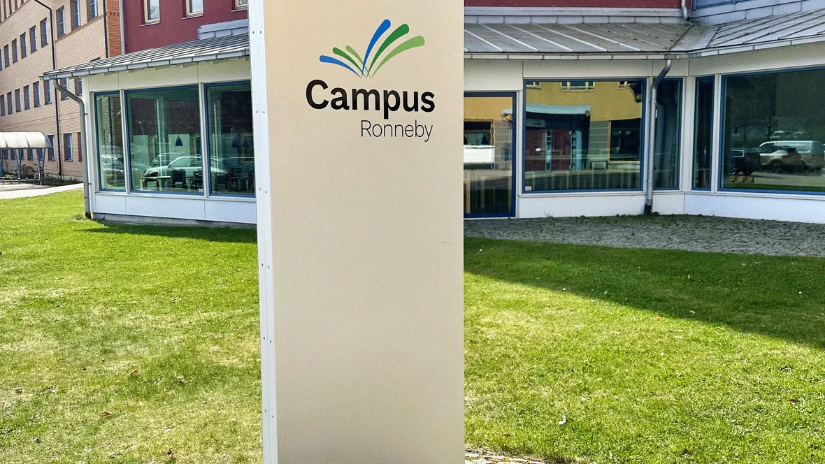 En stående rektangulär skylt med texten ”Campus Ronneby” och en grafisk logotyp i grönt och blått. Skylten är placerad på en stenbelagd yta framför en modern byggnad med röd och beige fasad och flera små fönster. Gräsmatta syns i bakgrunden.