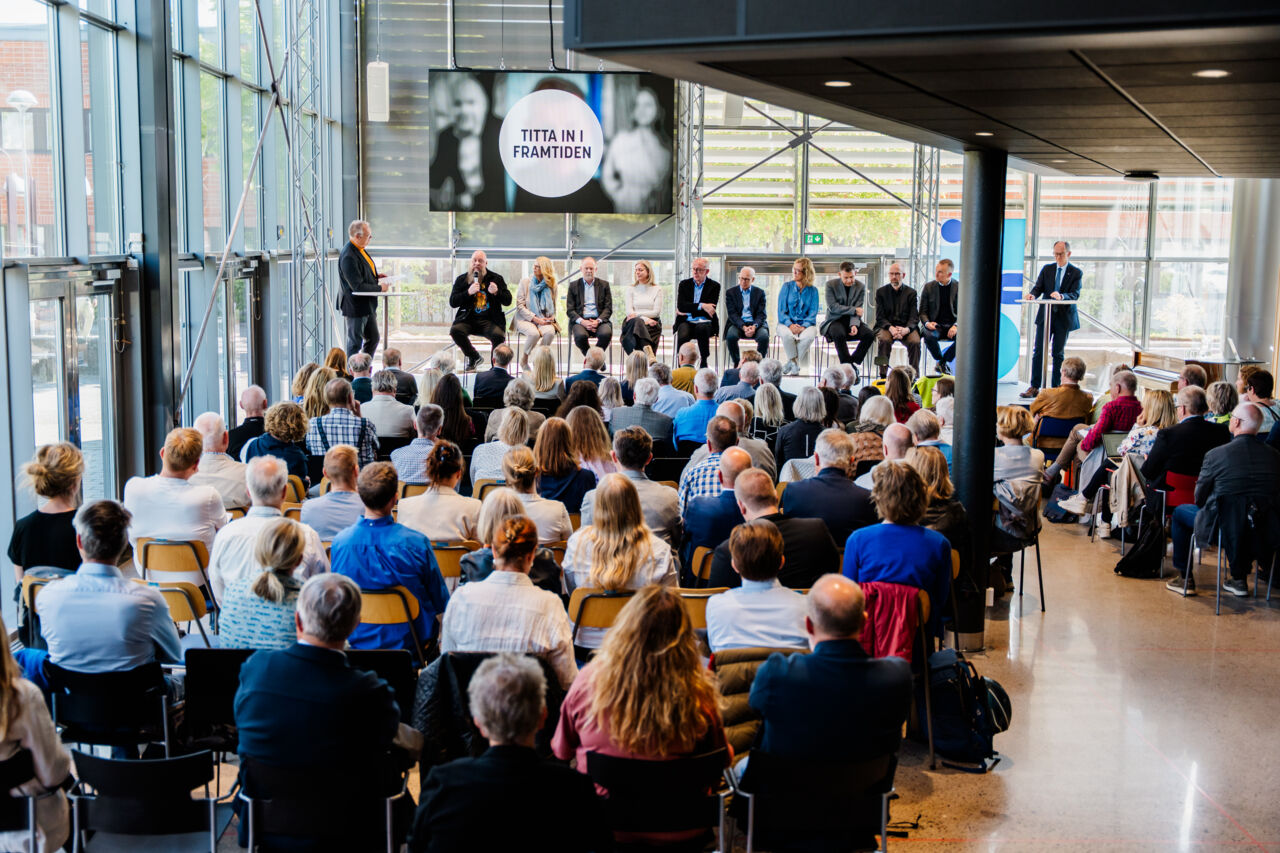 Publik bakifrån vänd mot en scen där människor sitter