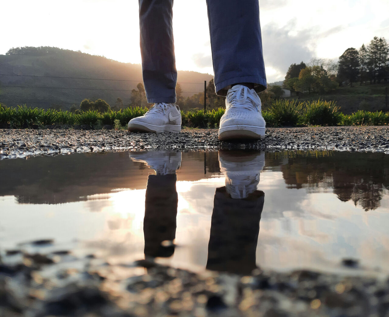A man is standing by a pool of water. His legs are reflected in the water.