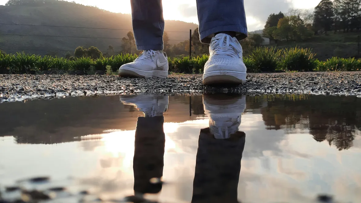 A man is standing by a pool of water. His legs are reflected in the water.