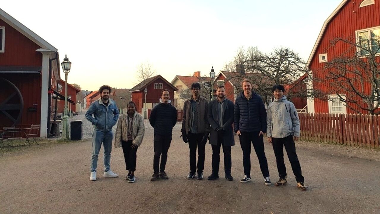 Seven people stand in a row on a dirt road lined with red wooden houses and leafless trees, facing the camera in daylight.