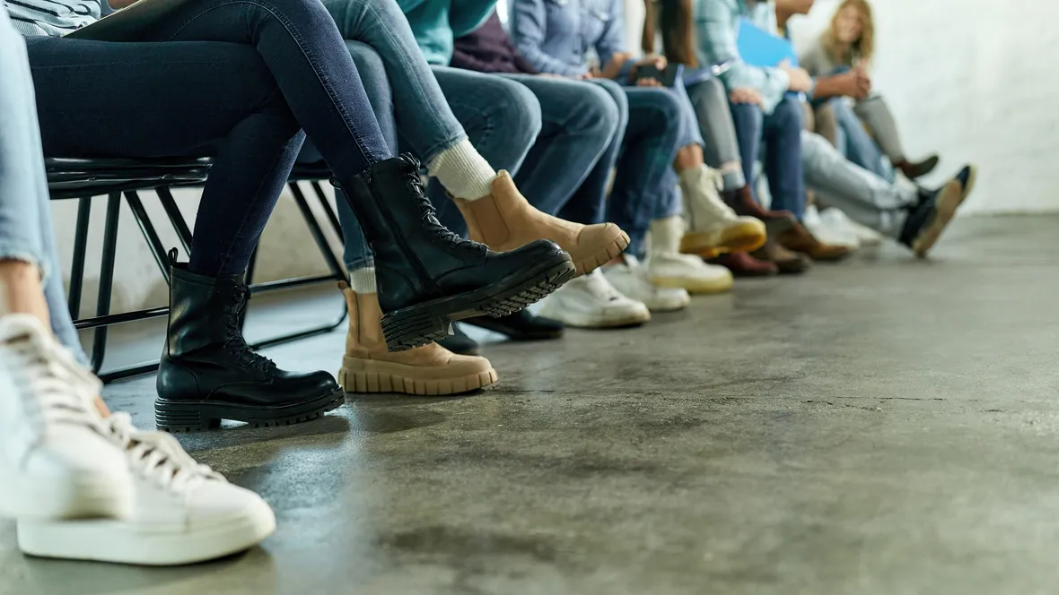 Människor som sitter på stolar i en lång rad, bilden visar endast ben och skor.