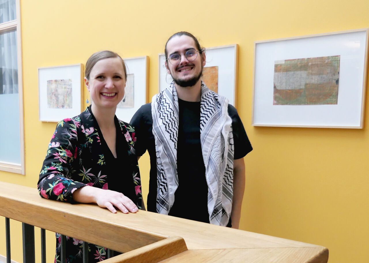 A man and a woman are standing in a corridor in front of a yellow wall with framed art. Both are smiling at the camera. The man, with dark hair, a beard, and glasses, has a keffiyeh draped over his shoulders. The woman, with her blonde hair in a bun, is leaning on a wooden railing and wearing a dark blouse with a colorful floral pattern.