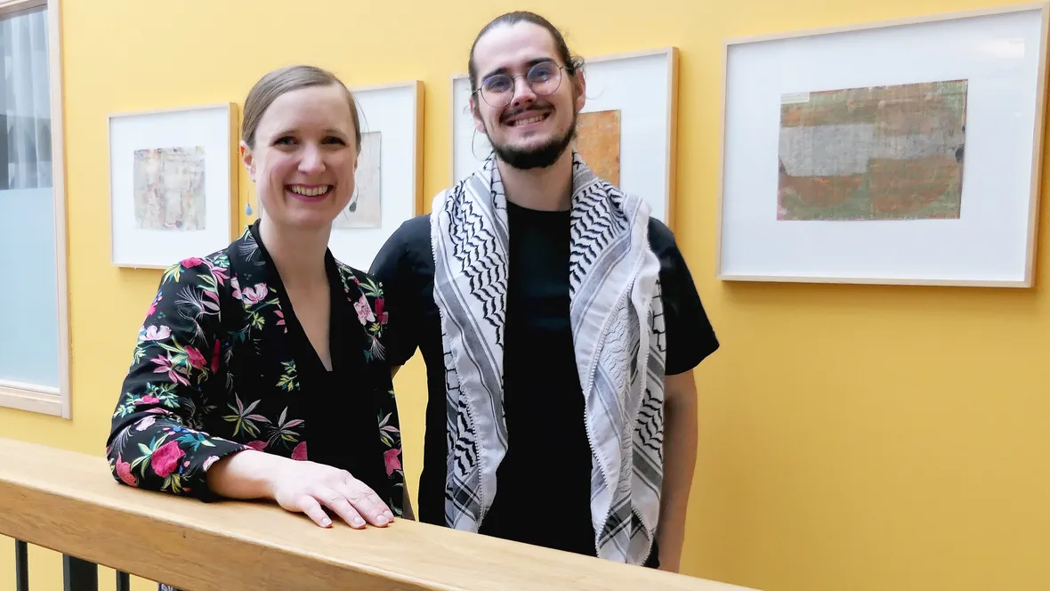 A man and a woman are standing in a corridor in front of a yellow wall with framed art. Both are smiling at the camera. The man, with dark hair, a beard, and glasses, has a keffiyeh draped over his shoulders. The woman, with her blonde hair in a bun, is leaning on a wooden railing and wearing a dark blouse with a colorful floral pattern.