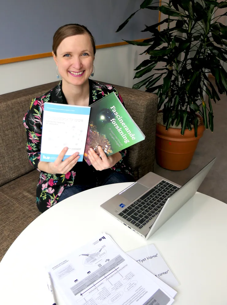 A woman with a smile on her face is seated on a sofa, holding up two books. One of the books is titled 