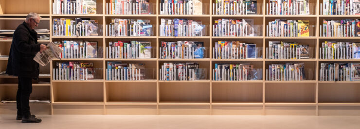 A shelf with journals and a man standing and reading a news paper.