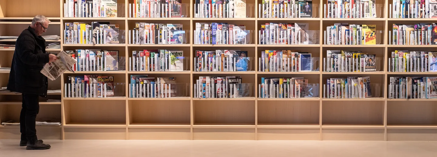 A shelf with journals and a man standing and reading a news paper.