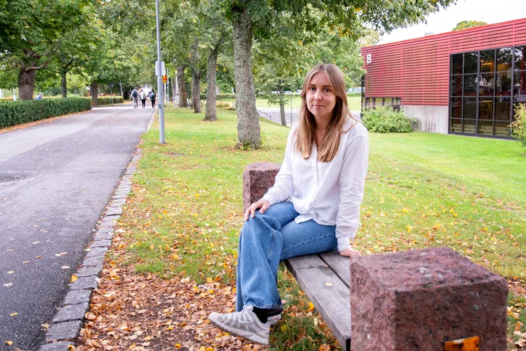 Katarina Soldan sitter på en bänk utomhus längs en asfalterad väg, träd och B-huset syns delvis i bakgrunden. 