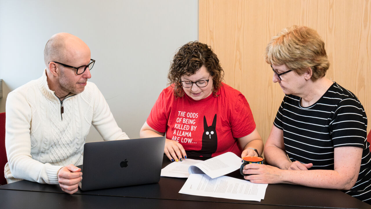 Tre personer sitter vid ett svart bord, på bordet finns ett dokument och en dator.