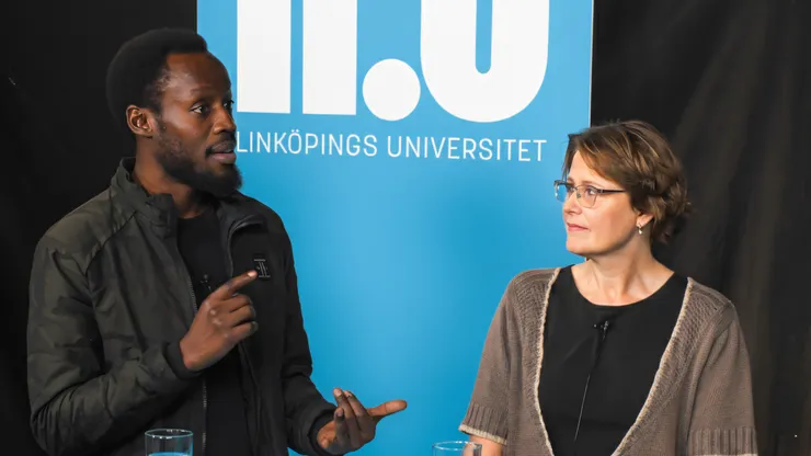 Two people are engaged in a discussion in front of a blue banner with the text ‘Linköpings universitet’. The man on the left, wearing a dark jacket, is gesturing with his hand while speaking. The woman on the right, wearing glasses and a brown knitted cardigan, is looking at him attentively. Both have microphones attached to their clothing.