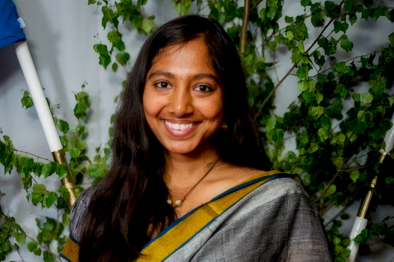 A close-up portrait of a smiling woman with long, dark hair. She is wearing a traditional garment in grey and yellow and a necklace with a pearl pendant. Lush green leaves and branches are visible in the background.