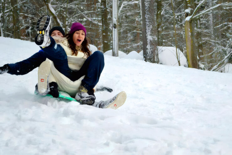 Two people are sledding down a snowy slope in a forest. The person in the background, wearing a purple hat, white jacket, and dark pants, is looking forward with an open-mouthed, surprised expression. The person in the foreground, only partially visible, is lying on their back with one boot lifted into the air. Both are dressed in winter clothing. The setting is a snow-covered forest.