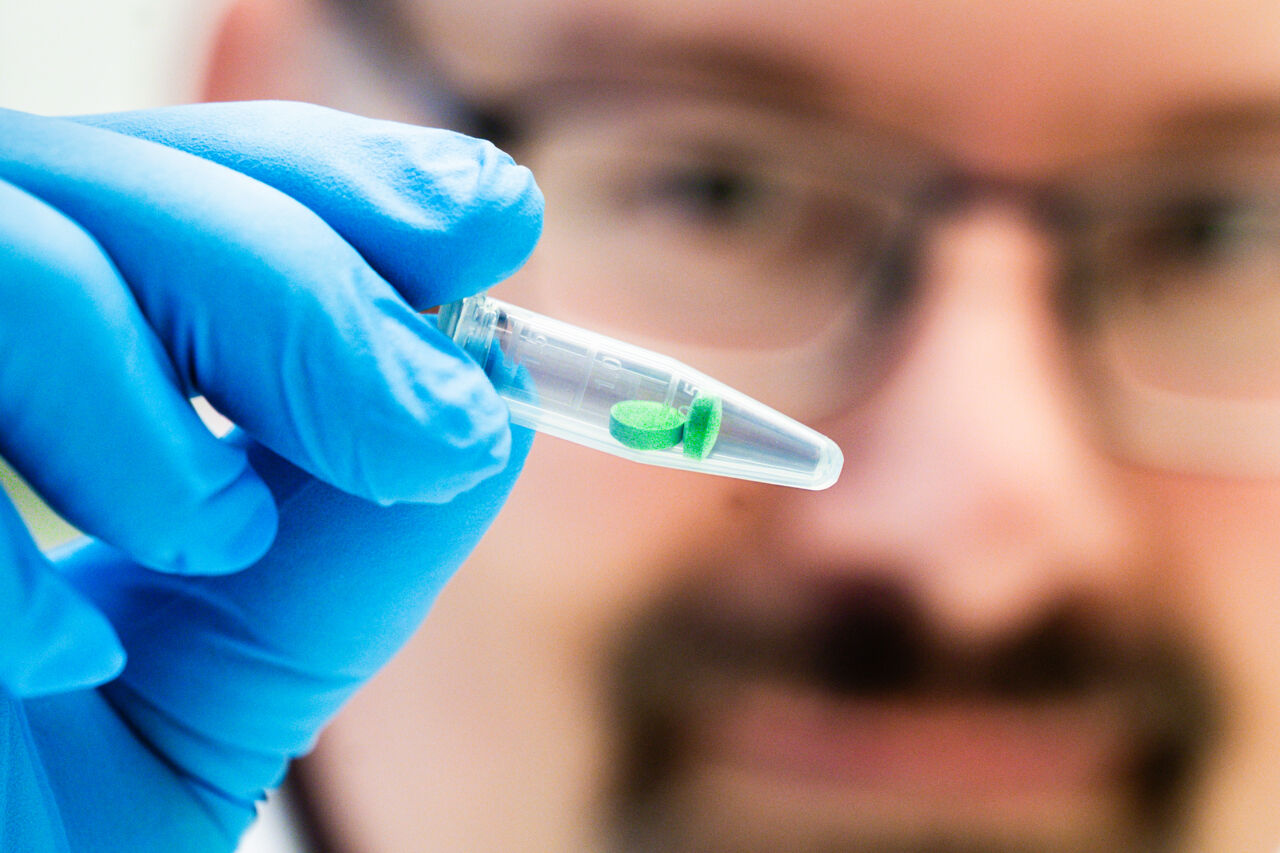 Man wearing a lab coat holds a test tube containing two green pills.