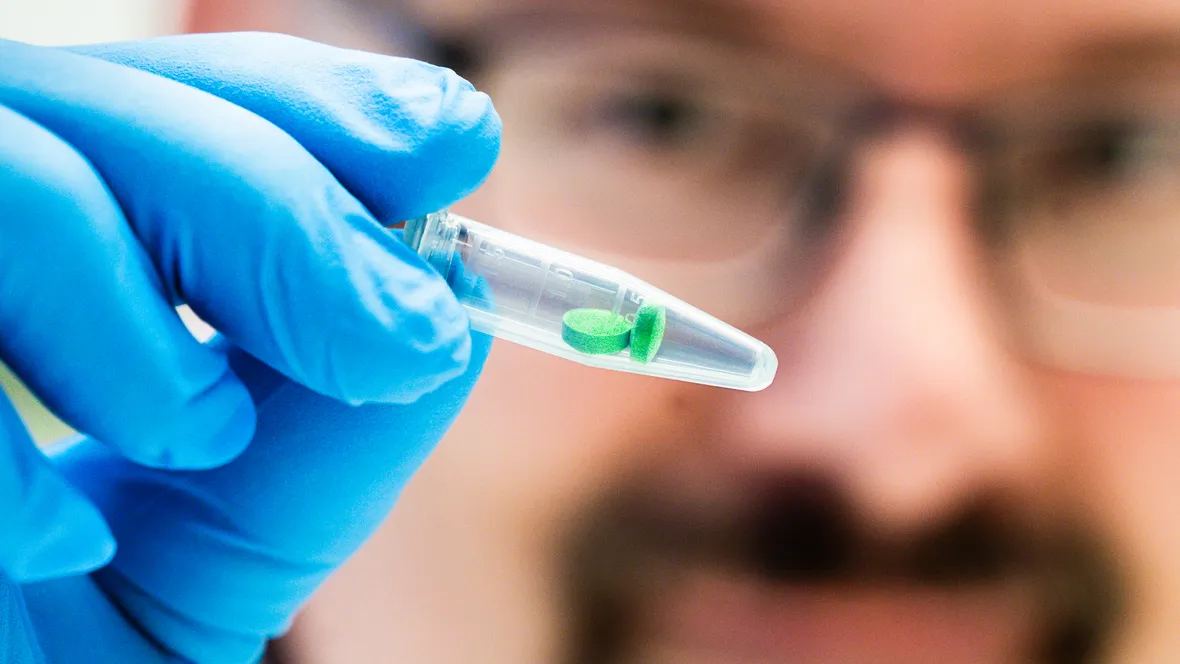 Man i labbrock håller upp provrör innehållandes två gröna tabletter.