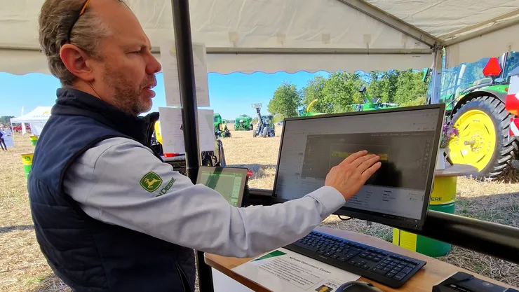 En man i en John Deere-väst använder en pekskärmsmonitor i en utomhusmonter med John Deere-branding, och jordbruksutrustning syns i bakgrunden.