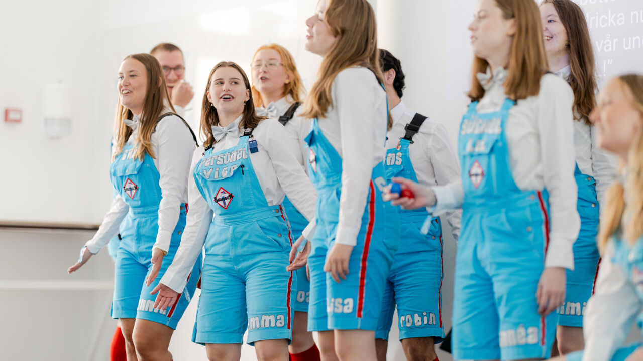 Nio studenter i knallblå kortbyxor och röda knästrumpor uppträder framför en whiteboard.