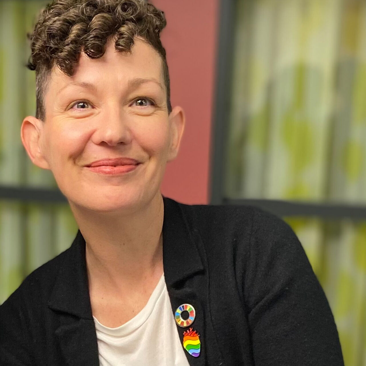 Person med kort lockigt hår ler mot kameran i inomhusmiljö.