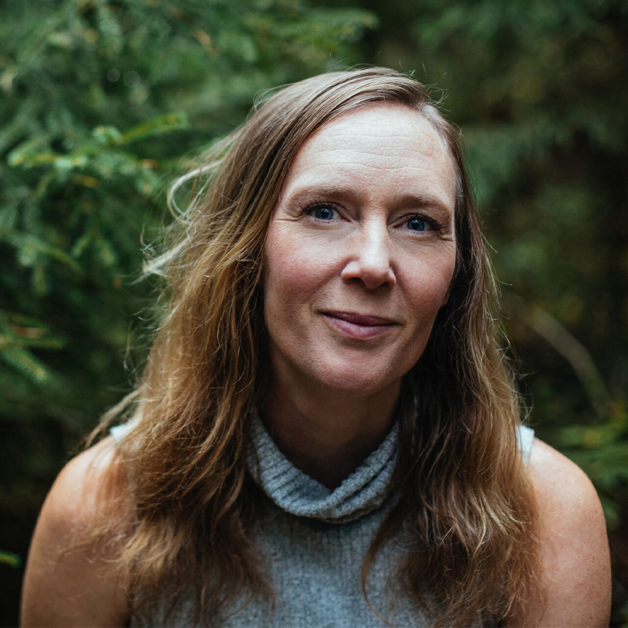 Person med långt hår ler försiktigt i en skogsmiljö.