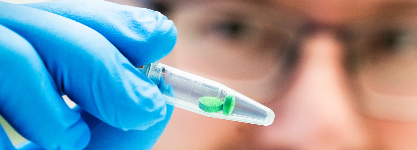 Man i labbrock håller upp provrör innehållandes två gröna tabletter.