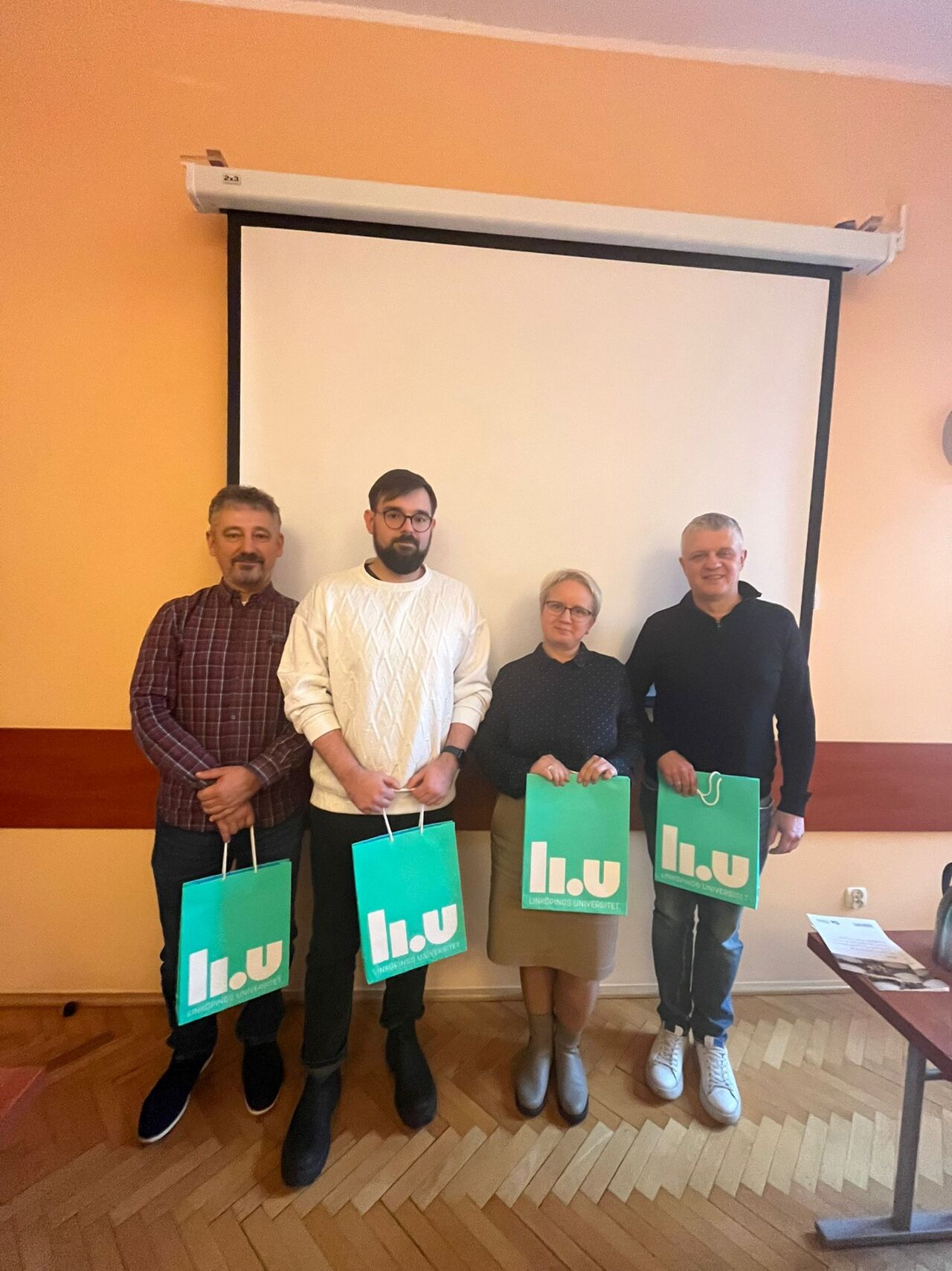 Four people standing in front of a screen and holding one green paper bag each
