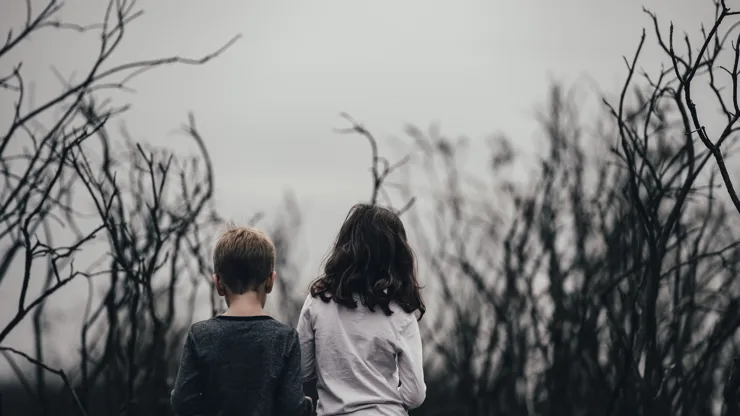 Två barn står med ryggen mot kameran bland torra, nakna grenar under en grå himmel.
