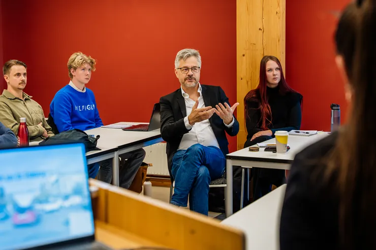 A man is sitting and talking in a lecture hall with students around him.