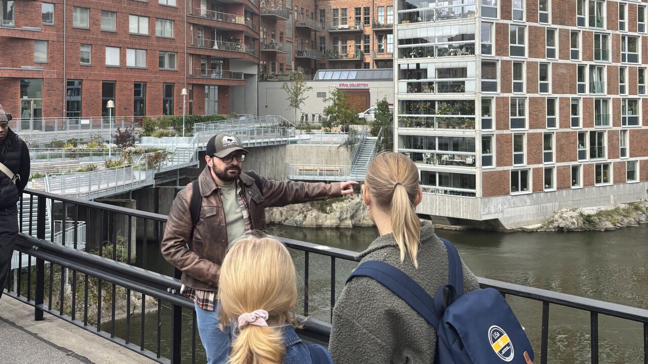 En man står utomhus på en bro och talar inför flera personer.