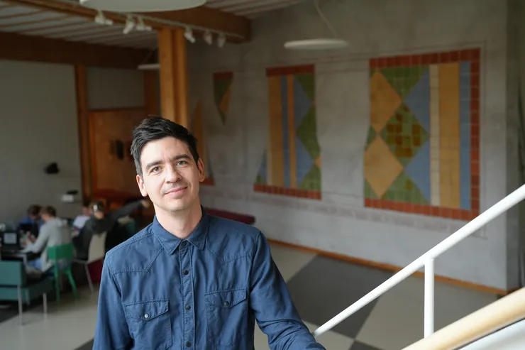 A person is standing indoors on a staircase with people blurred in the background.