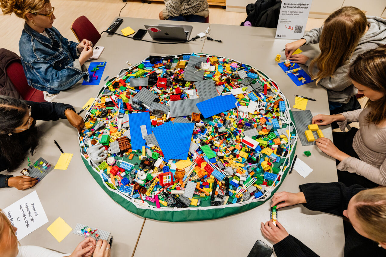 Massor av legobitar i  en stor hög på ett bord med fler personer runt som bygger med lego.