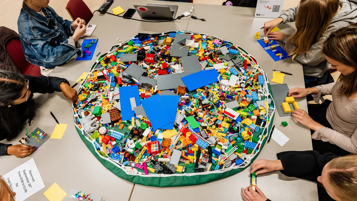 Lots of lego pieces in a big pile on a table, with several people around building with lego