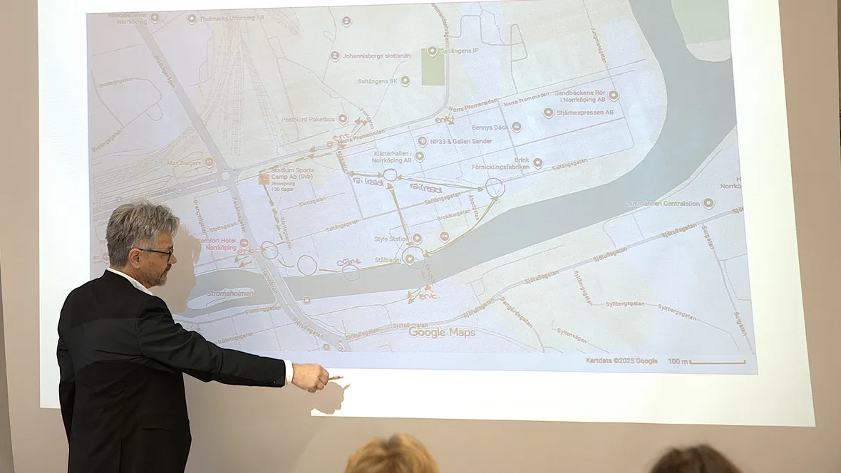 A person is standing in a lecture hall by a projection screen and pointing at a map.